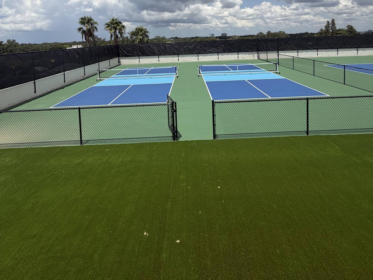 Two fenced rooftop pickleball courts beside a turf field, with palms and clouds beyond