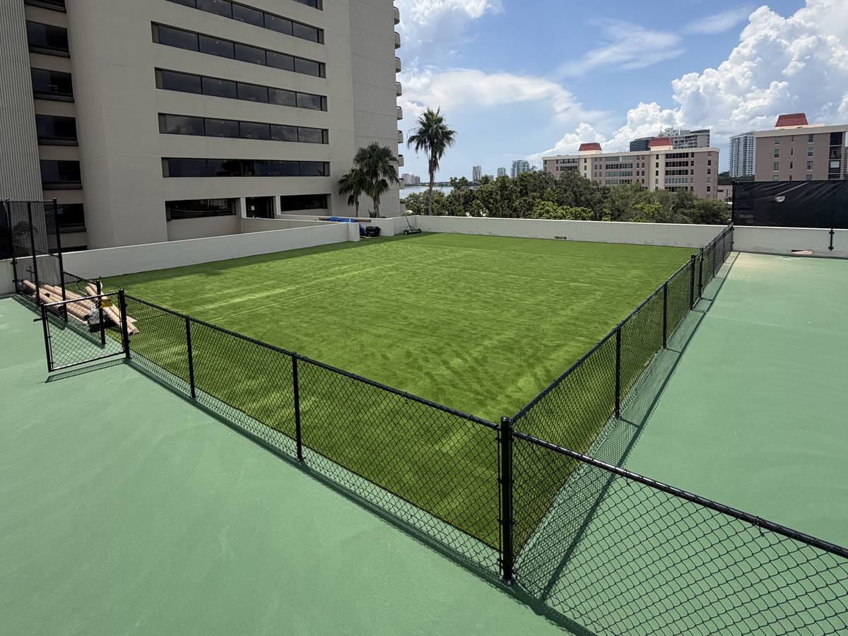 Rooftop turf field inside a black chain-link fence beside a high-rise tower, with palms and the skyline beyond