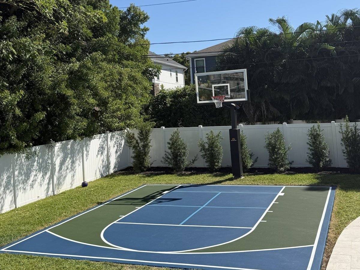 Backyard basketball court in blue and green with a white vinyl fence, hedges and a glass-backboard goal